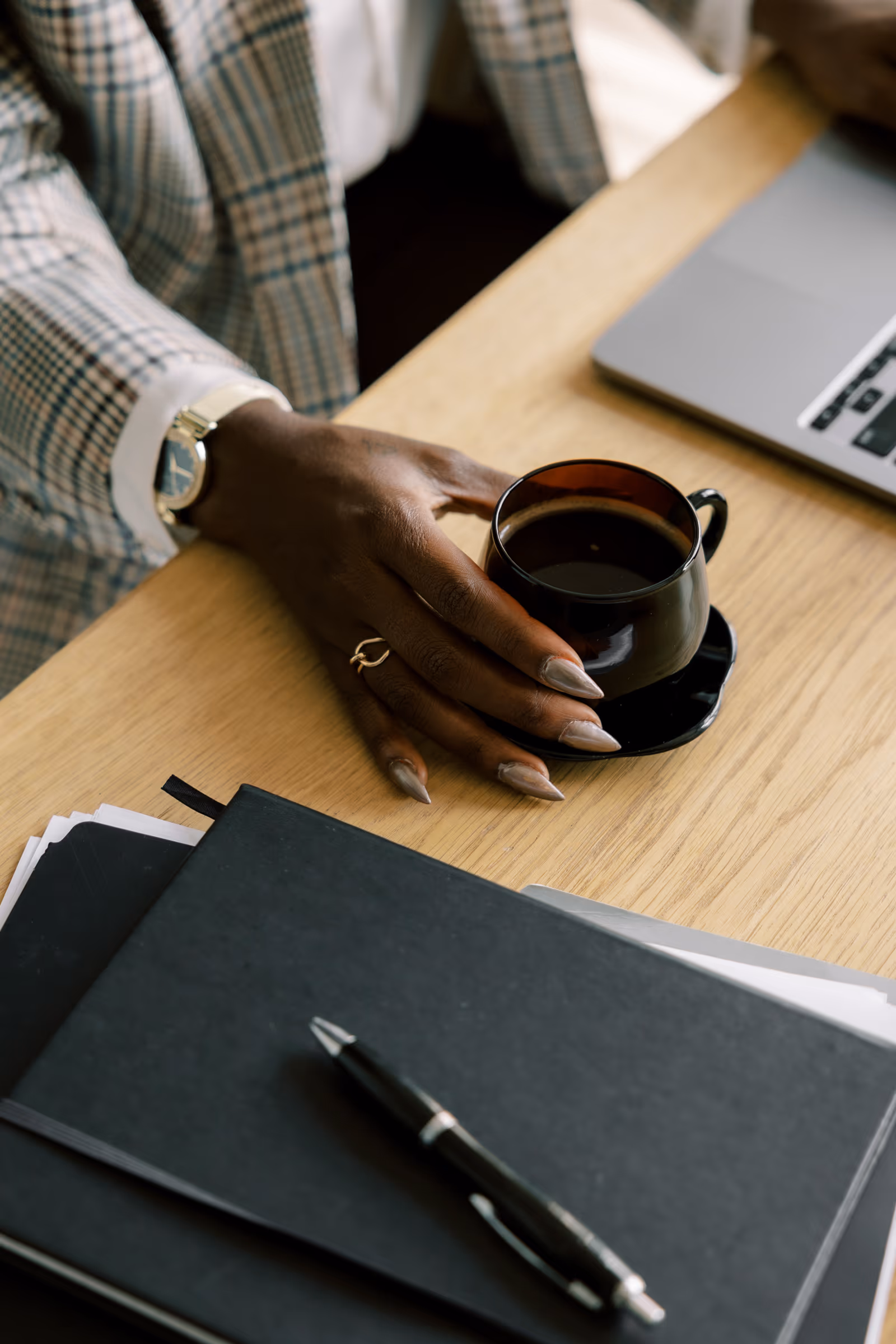 Close-up of a woman holding a cup of coffee beside a notebook and laptop.