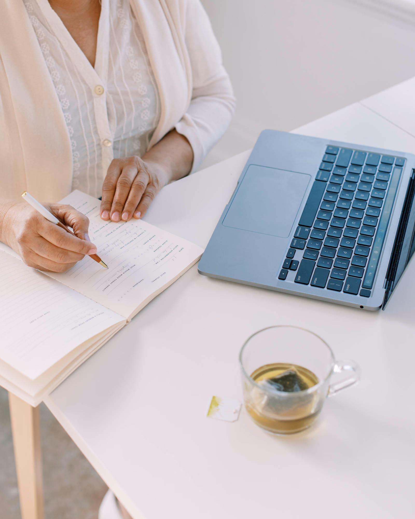 Woman writing in a notebook beside a laptop at a desk.